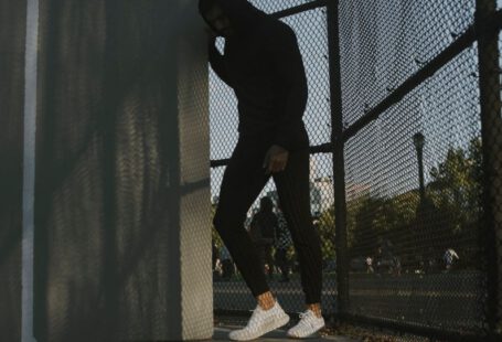 Athletic Shoes - a man in a black hoodie leaning against a fence