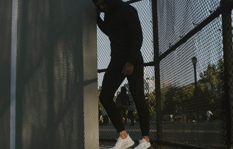 Athletic Shoes - a man in a black hoodie leaning against a fence